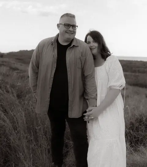 Black-and-white portrait of a couple standing together in coastal grass.